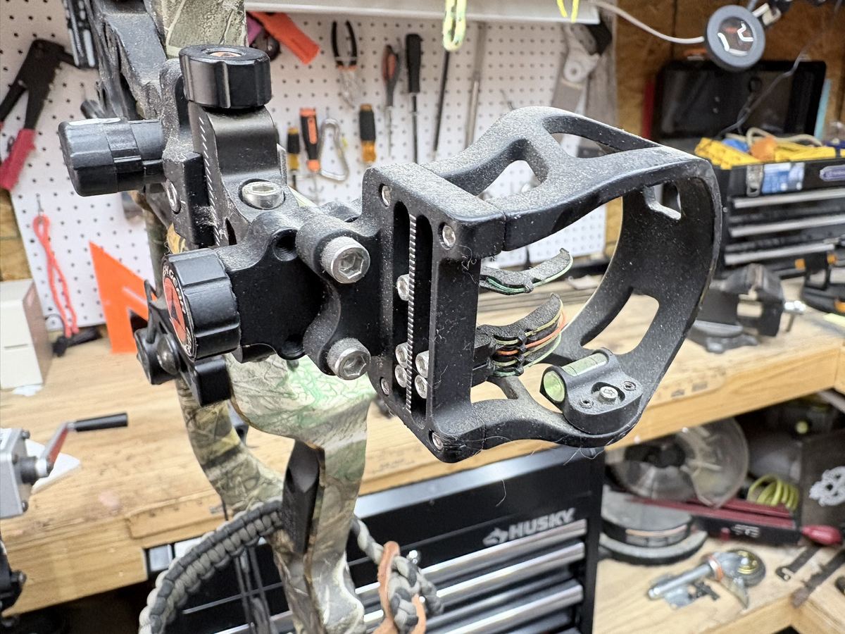 A multi-pin bow sight with a bubble level mounted on a camo compound bow in the shop