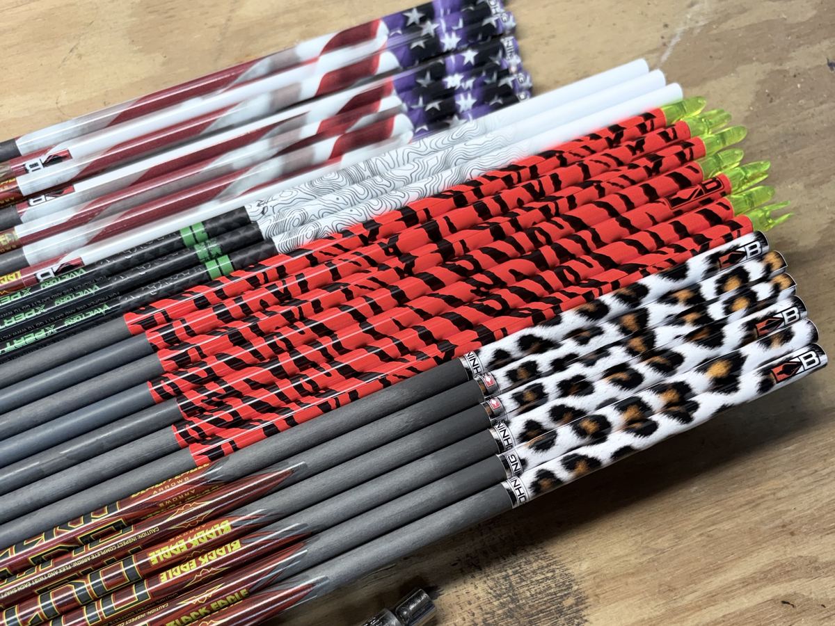 A row of carbon arrows with flag, tiger-stripe and leopard-print wraps laid out on a workbench