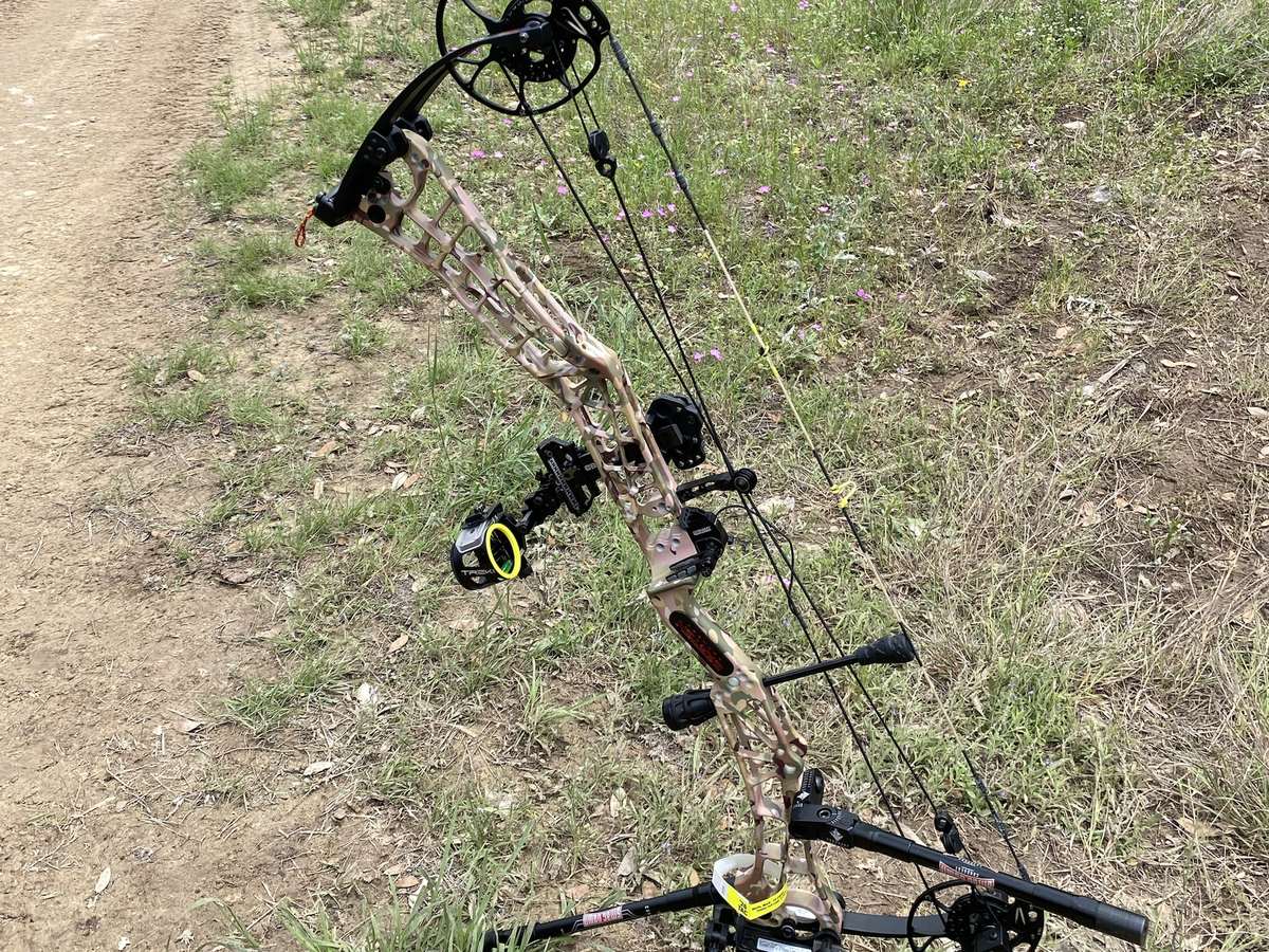 A camo compound bow with a sight, arrow rest and stabilizer installed, on a bow stand outdoors